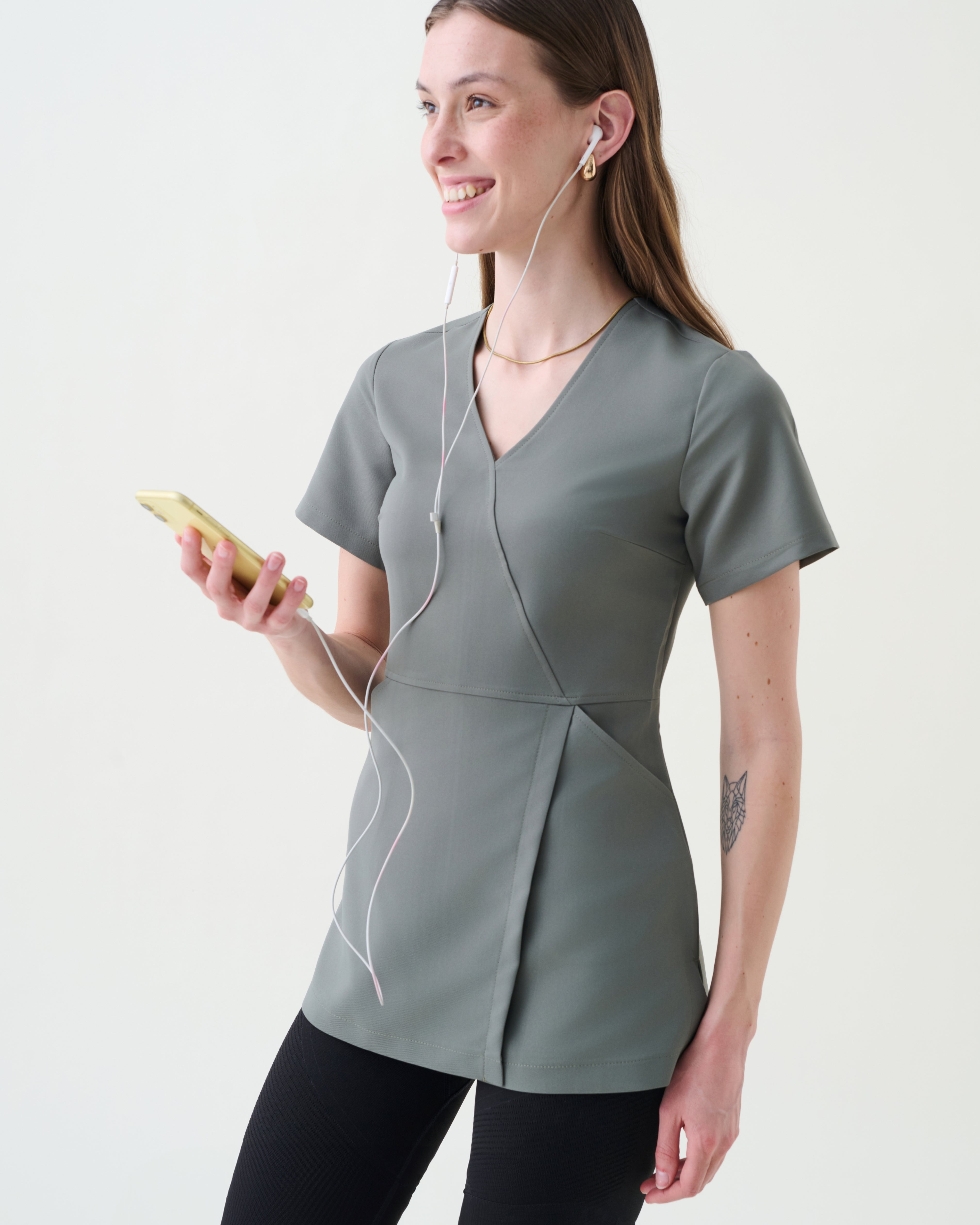 Woman wearing a gray top and black pants, holding a phone and earphones, on a white background