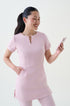 A woman standing against a white background wearing a blush pink beauty and clinic tunic with front pockets and matching pants with her phone.