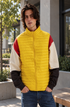 A man wearing a bright yellow quilted puffer vest with color-blocked sleeves in red, cream, and black, paired with blue jeans, standing outdoors in front of a modern building.