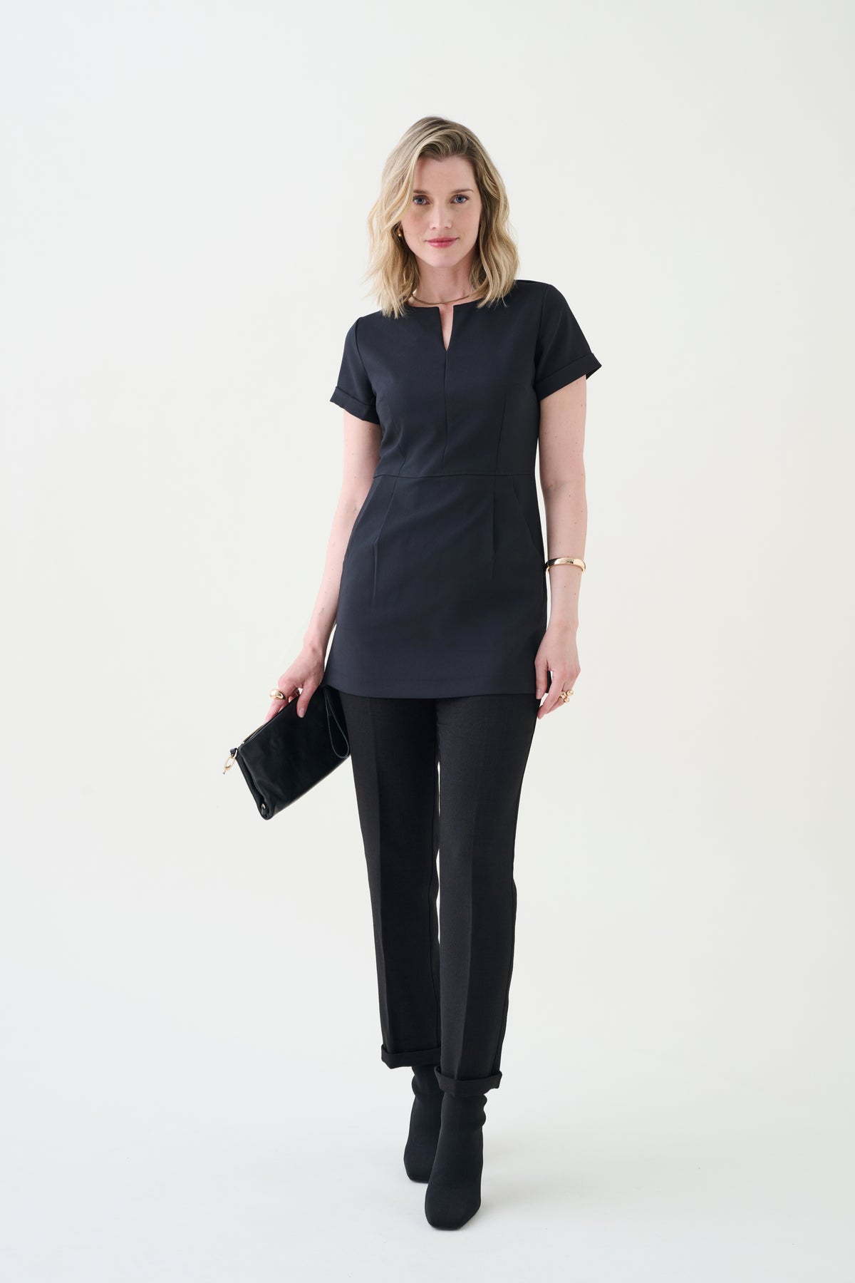 A woman standing against a white background wearing a beauty and hotel black tunic with front pockets and matching black pants.